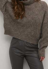 Gruby beżowo-brązowy sweter z golfem, wykonany w ażurowy wzór z ściągaczami na mankietach, zestawiony z ciemnoszarymi spodniami z ekoskóry.