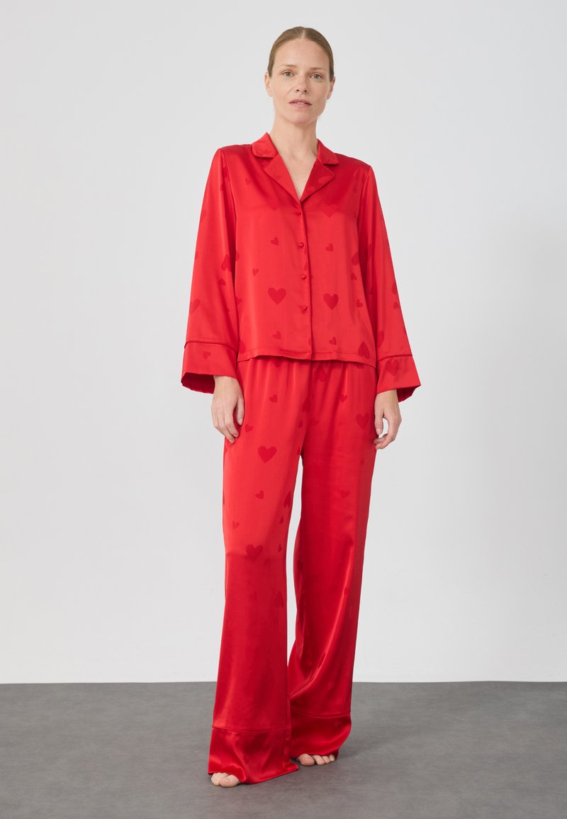 Set di pigiama in satin rosso con motivi a cuore, composto da una camicia con colletto e bottoni e pantaloni a gamba larga. Tessuto morbido e vestibilità rilassata.