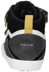 Zapato de ante negro y gris hasta el tobillo con una suela de goma blanca, que presenta una correa amarilla y un logotipo en la parte posterior. Cierre de cremallera en el lateral.