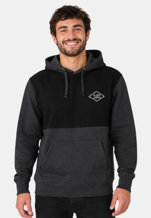 Hombre sonriente con sudadera con capucha negra y gris oscuro con el logo "Lee Cooper" en el pecho, de pie frente a un fondo claro y liso.