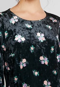 Svart blommig klänning med ett mjukt, texturerat tyg. Rund halsringning med små mönster av rosor och lila blommor. Löst sittande med långa ärmar.