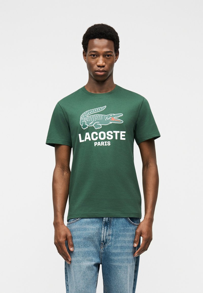 Νεαρός άνδρας που φοράει πράσινο μπλουζάκι Lacoste Paris με λευκό λογότυπο κροκοδείλου και ανοιχτόχρωμο τζιν, στέκεται μπροστά σε λευκό φόντο.