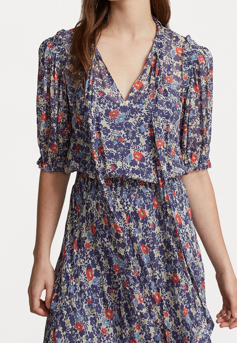Femme portant une robe fleurie avec des manches bouffantes, un col en V et une taille cintrée, avec des fleurs rouges, bleues et violettes sur un fond clair.