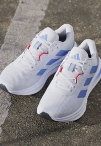 Baskets blanches en matière mesh, avec des accents bleu clair et rouge, et une surface texturée. La semelle est grise et de forme arrondie.