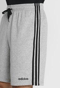 Grå atletiska shorts med dragsko i midjan, prydda med tre svarta vertikala ränder längs sidorna och en Adidas-logga längst ner till vänster.