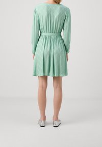 Vrouw in een mintgroene jurk tot de knie met lange mouwen en een riem, staand met de rug naar ons gekeerd, vergezeld van zilveren platte schoenen op een effen achtergrond.