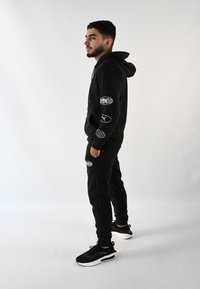 Conjunto de sudadera con capucha y pantalones de chándal negros con logotipos, que presenta un corte relajado y capucha con cordón. Zapatillas negras con suelas blancas.