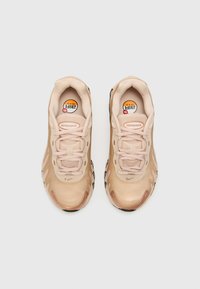 Beige Nike Air Max sneakers hebben een gladde, glanzende bovenkant, vetersluiting en een zwarte rubberen buitenzool met ribbels. "Air Max" branding is zichtbaar.