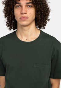 Mørkegrøn kortærmet bomulds t-shirt med rund hals og en brystlomme. Stoffet er glat med en ensartet struktur.