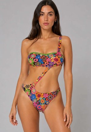 Donna che indossa un costume da bagno monospalla con taglio asimmetrico e motivo floreale colorato all'uncinetto, in piedi davanti a uno sfondo grigio uniforme.