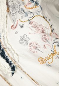 Bílá látka s barevným květinovým vyšíváním v růžových, modrých a kovově stříbrných nitích. Text "Desigual" je výrazně zobrazen. Texturované okraje.