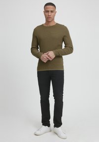 Olivengrønn ribbestrikket genser med lange ermer, rund hals, kombinert med svarte jeans og hvite sneakers; synlig logo detaljer på genserens kant.
