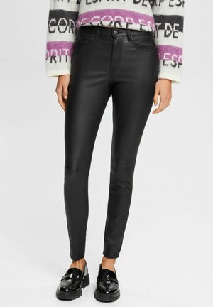 Woman wearing black slim-fit leather pants, a white and purple patterned sweater, and black shiny loafers, standing against a plain background.