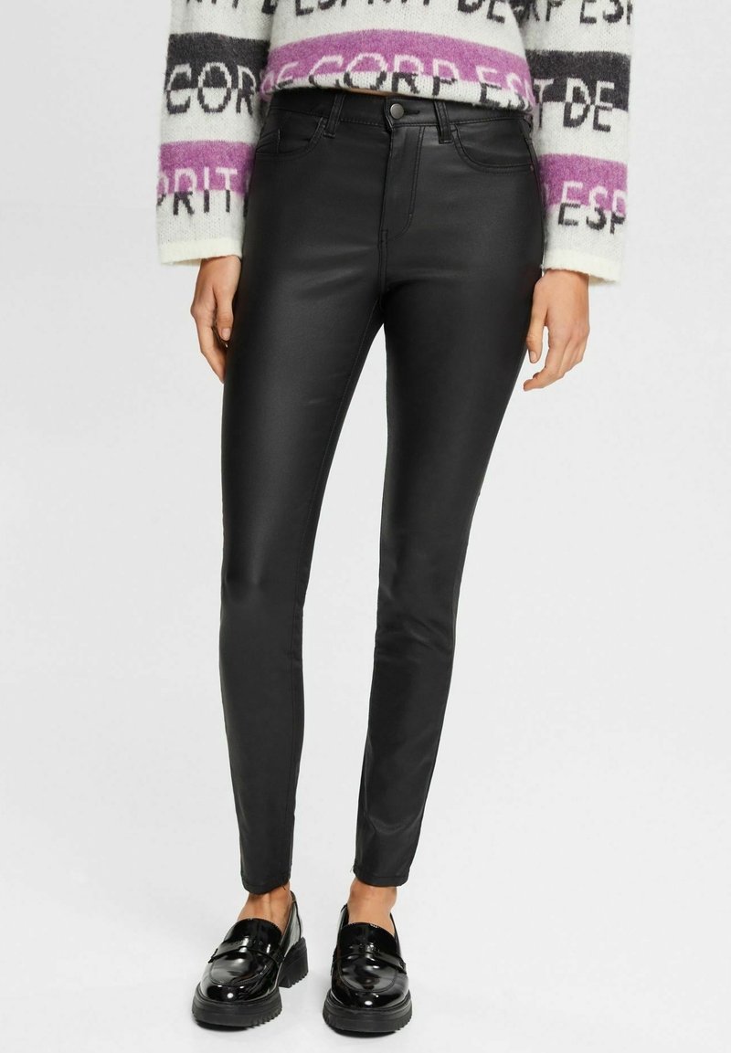 Femme portant un pantalon en cuir noir ajusté, un pull blanc à motifs violets, et des mocassins noirs brillants, debout devant un fond uni.