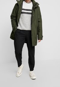 Oliwkowa kurtka parka na guziki, szary sweter dziany z czarnymi paskami, czarne spodnie i białe trampki z czerwonymi akcentami.