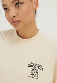 Junge Frau trägt ein cremefarbenes T-Shirt mit schwarzem Aufdruck "Always Stay Groovy DISCOTECA" und einer Diskokugel-Grafik auf der Brust, silberne Kreolen.