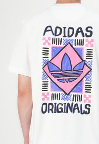 Hvit bomull T-skjorte med et fargerikt grafisk design på baksiden, inkludert en rosa diamantform med en blå Adidas-logo og fet tekst.