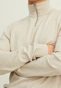 Pull beige à fermeture éclair avec col montant, poignets côtelés et texture lisse. Dispose d'une fermeture éclair argentée.