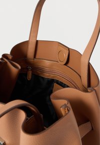 Sac fourre-tout en cuir fauve avec une surface texturée, un dessus ouvert et une poche intérieure zippée. Comprend des anses lisses et une doublure en tissu noir.