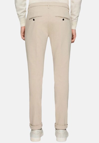 Pantaloni beige su misura con taglio dritto, dotati di tasche posteriori con bottoni neri e risvolti. Realizzati in un tessuto liscio, abbinati a sneakers bianche.