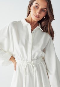 Abito bianco con bottoni, vita regolabile con cordino, maniche lunghe con polsini elastici e un tessuto liscio e testurizzato. Orecchini dorati in vista.
