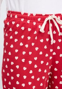 Κόκκινο παντελόνι πιτζάμας με ελαστική μέση και λευκό κορδόνι. Διακοσμημένο με λευκά σ χέδια καρδιών ομοιόμορφα κατανεμημένα σε όλο το ύφασμα.