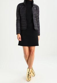 Veste noire matelassée avec un col montant, associée à une robe en tricot noir à côtes et des bottines dorées brillantes, mettant en valeur une silhouette élégante.