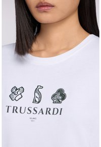 Biała koszulka z krótkim rękawem z czarnymi graficznymi wzorami logo: dłoń, pies i koniczyna, z napisem "TRUSSARDI MILANO 1911" poniżej.
