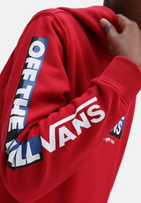 Sudadera roja con capucha y puños acanalados, presenta un llamativo texto "OFF THE WALL" en azul y blanco en las mangas. Material de algodón, acabado texturizado.