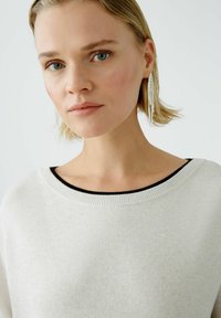 Hellgrauer Pullover mit gerippter Textur, weit geschnittenem Halsauschnitt mit schwarzem Akzent. Das Model trägt lange, silberne, hängende Ohrringe.