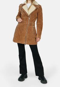 Chaqueta de ante marrón con cuello forrado de borrego, bolsillos con cremallera, cintura con cinturón y detalles en metal plateado.