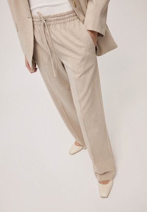 Frau trägt beige Kordelzug-Hosen kombiniert mit einem hellbeigen Blazer und weißen Schuhen mit quadratischer Spitze vor einem schlichten Hintergrund.
