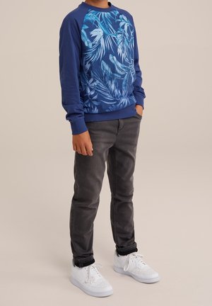 Kind trägt ein blaues Langarmshirt mit hellblauen Blattmustern, dunkelgraue Hose und weiße Turnschuhe, steht mit einer Hand in der Tasche.