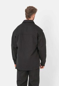 Chemise-veste noire, surdimensionnée, avec un col, des manches longues et une texture lisse. Le dos présente un détail de couture et des courbes d'ourlet subtiles.