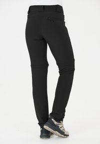 Pantalons de randonnée noirs avec un design ajusté, poche zippée à l'arrière et tissu extensible ; portés avec des chaussures de randonnée grises et beige.