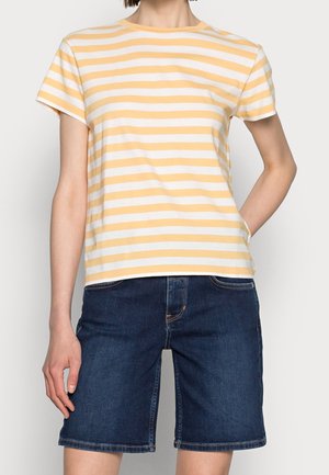 Persoon die een geel-wit gestreept korteme T-shirt draagt met donkerblauwe denim knielange shorts, staand met één hand in de zak.