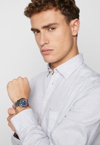 Montre en argent sur un bracelet en maille métallique, avec un cadran bleu agrémenté d'accents rouges et blancs, portée avec une chemise boutonée gris clair.