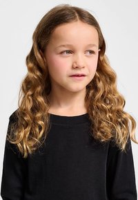 Czarna koszula z długim rękawem o prążkowanej fakturze. Osoba ma falowane, sięgające ramion włosy koloru karmelowego oraz niebieskie oczy.