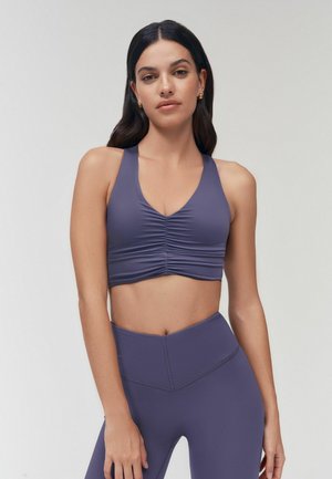 Kvinde med langt mørkt hår iført rynket lilla sports-bh og matchende højtsiddende leggings, stående mod en ensfarvet lys baggrund.