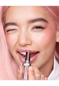 Transparent lipstick tube with bronze accent held close to lips. Model has light pink hair and a glossy, nude lip color. Smooth skin texture.