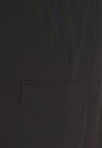 Czarna tkanina o gładkiej fakturze, zbliżenie na kieszeń z klapką w szpic, w prostym, minimalistycznym stylu.