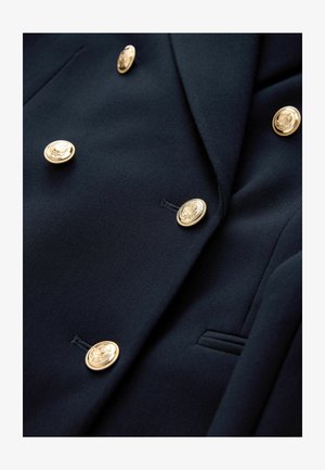 Navy blazer made of textured fabric with a double-breasted design, featuring six gold buttons with embossed detailing and a side pocket.