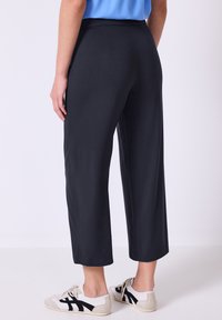 Pantaloni neri a cavallo corto con una texture liscia e vita elastica, abbinati a sneakers bianche e nere. Vista da dietro.
