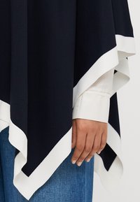 Námorní pončo s bílým lemováním, vyrobené z měkké látky. Má asymetrický lem a je nošeno přes bílé tričko a džíny.