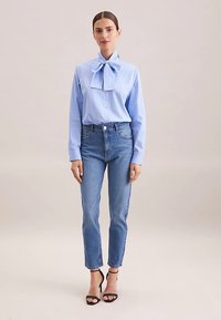 Camicia azzurro chiaro a righe sottili con un grande fiocco al collo, abbinata a jeans blu a vita alta e tacchi neri con cinturini.