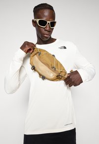 Brown waist bag with adjustable straps, zip closure, and front brand logo, paired with a white long-sleeve shirt.