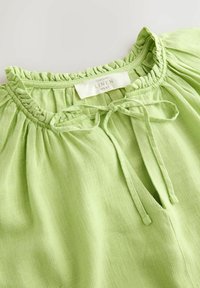 Haut en lin vert clair avec un col froncé. Présente un détail noué au niveau du cou et un tissu texturé avec un motif subtil.