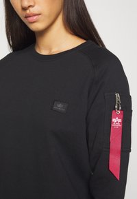 Sudadera negra con cuello de canalé, un parche con el logo en el pecho y un bolsillo con cremallera con una etiqueta de tela roja en la manga.