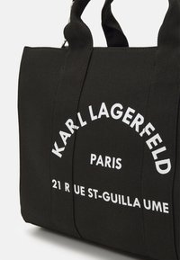 Mustat kangaskassit, joissa on kaksinkertaiset yläkahvat ja valkoinen teksti: "KARL LAGERFELD PARIS 21 RUE ST-GUILLAUME." Rakenteellinen muoto ja teksturoitu pinta.