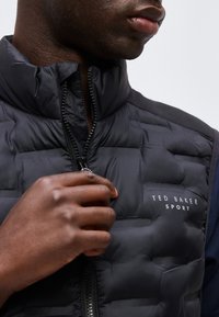 Άνδρας που κλείνει με φερμουάρ ένα μαύρο καπιτονέ μπουφάν Ted Baker Sport, εστιάζοντας στο χέρι, το φερμουάρ και το λογότυπο στο στήθος.